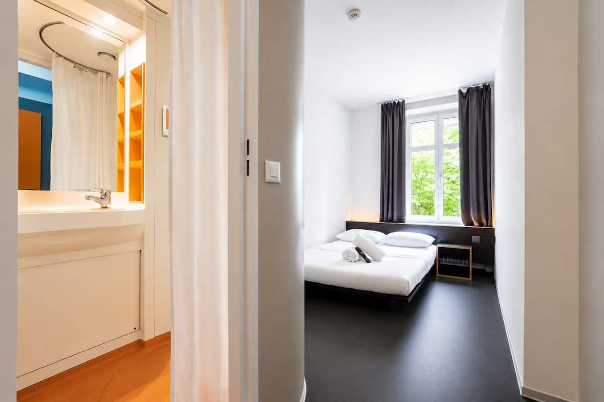 Una habitación de hotel con cama, ventana, mesita de noche y baño con lavamanos y espejo.