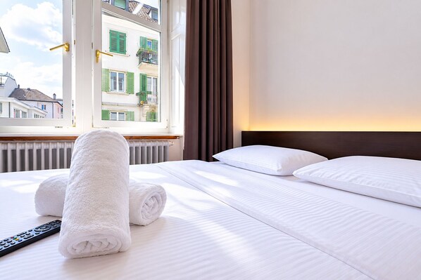 A neatly made bed with white linens and rolled towels, a remote control, and a window with a view of buildings.