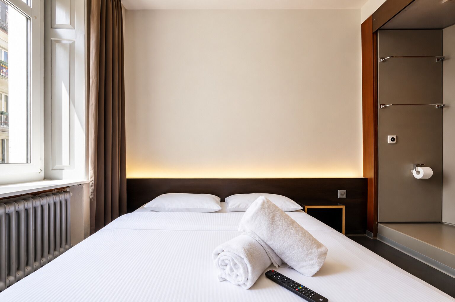A hotel room with a bed, a remote control, a towel, a window with curtains, and a door with a mirror.