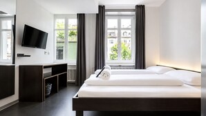 Una habitación de hotel moderna con una cama, un televisor montado en la pared, un escritorio y una ventana con cortinas.