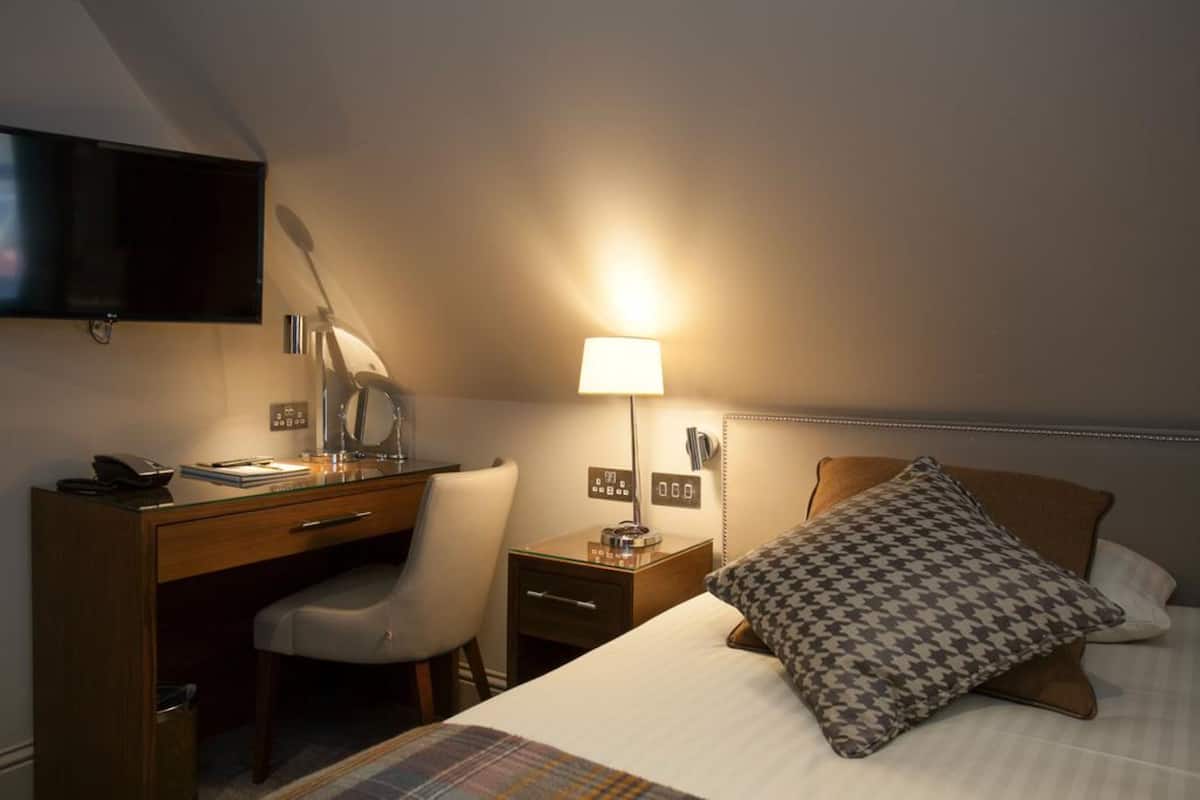 A hotel room with a desk, chair, bedside table, lamp, bed, and a television mounted on the wall.