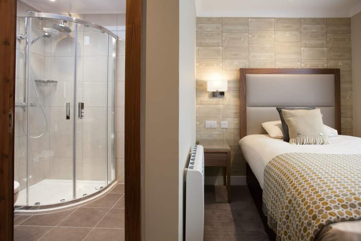A hotel room with a glass-enclosed shower, a bed with a headboard, and a nightstand with a lamp.