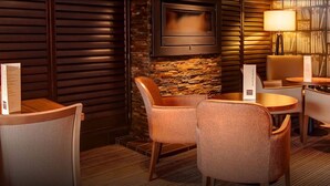 A cozy lounge area with a stone wall, wooden slats, and a bookshelf filled with books. There are several chairs and tables, and a floor lamp is visible. - Cruachan Hotel (Fort William)