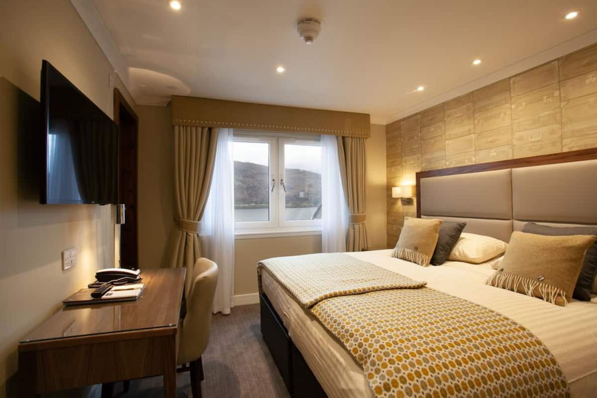 A hotel room with a large bed, a desk with a chair, a flat-screen TV, and a window with curtains.