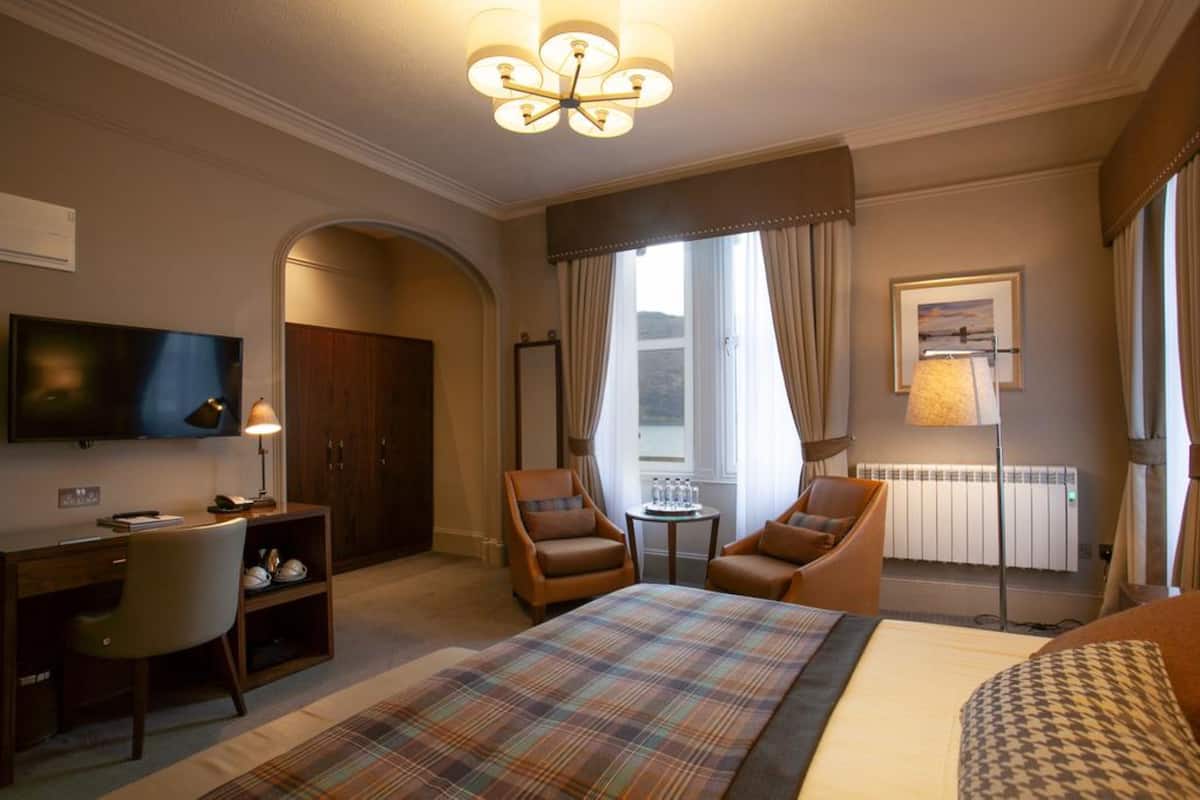 A hotel room with a bed, two armchairs, a desk, a television, a small table, and a window with curtains.