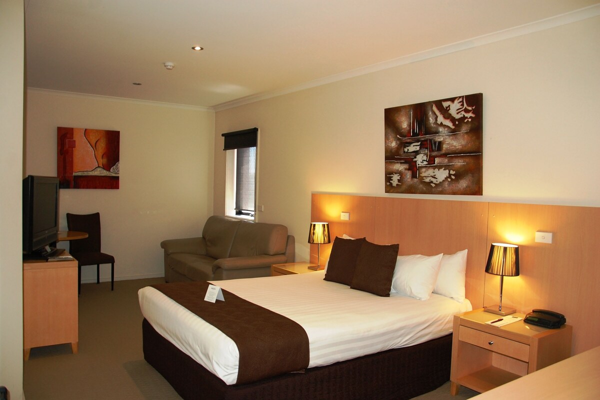 Habitación de hotel con una cama grande, lámparas de noche, un escritorio con lámpara, una silla, televisor y obras de arte en las paredes.