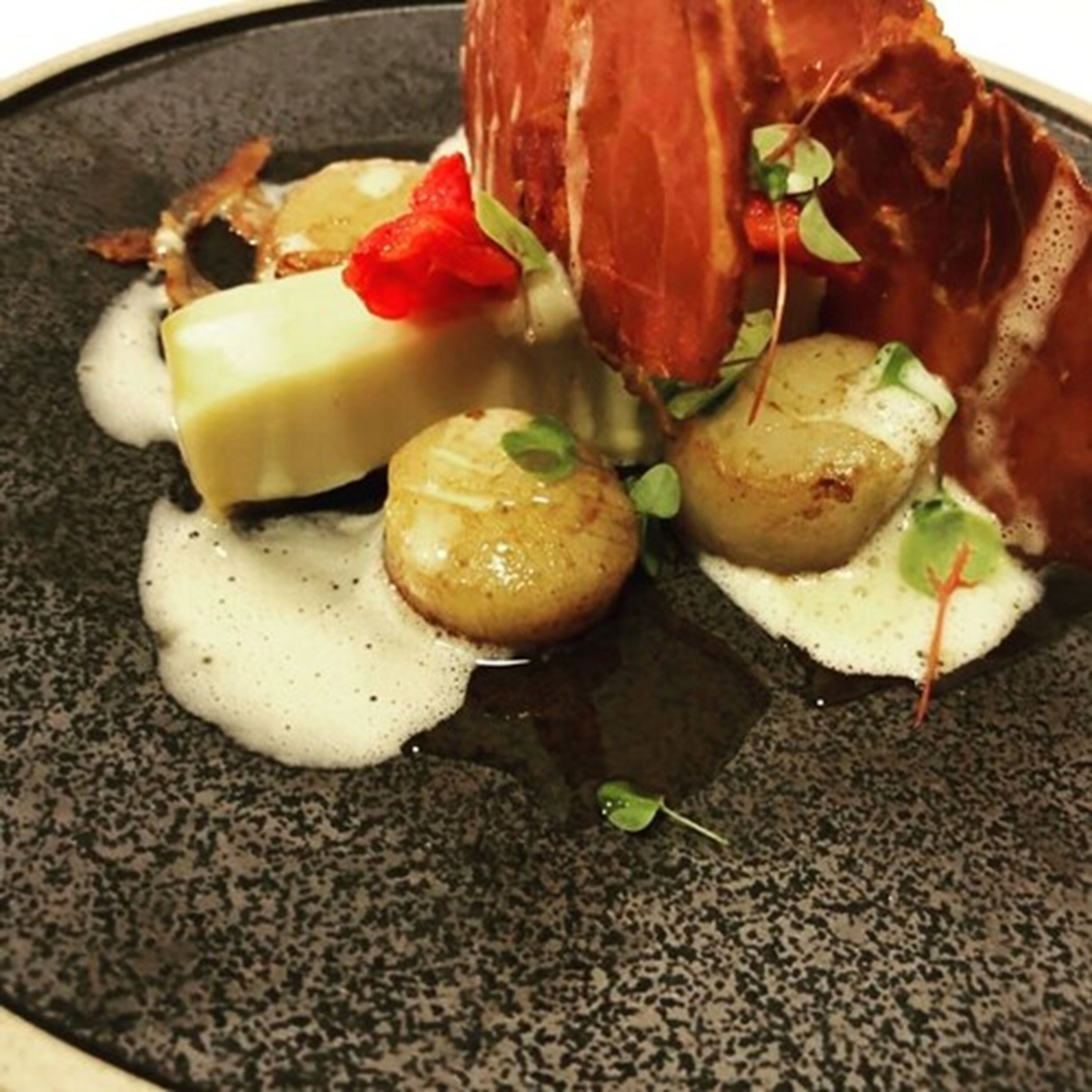 A dish with a piece of meat, a round vegetable, and a creamy sauce on a speckled plate.