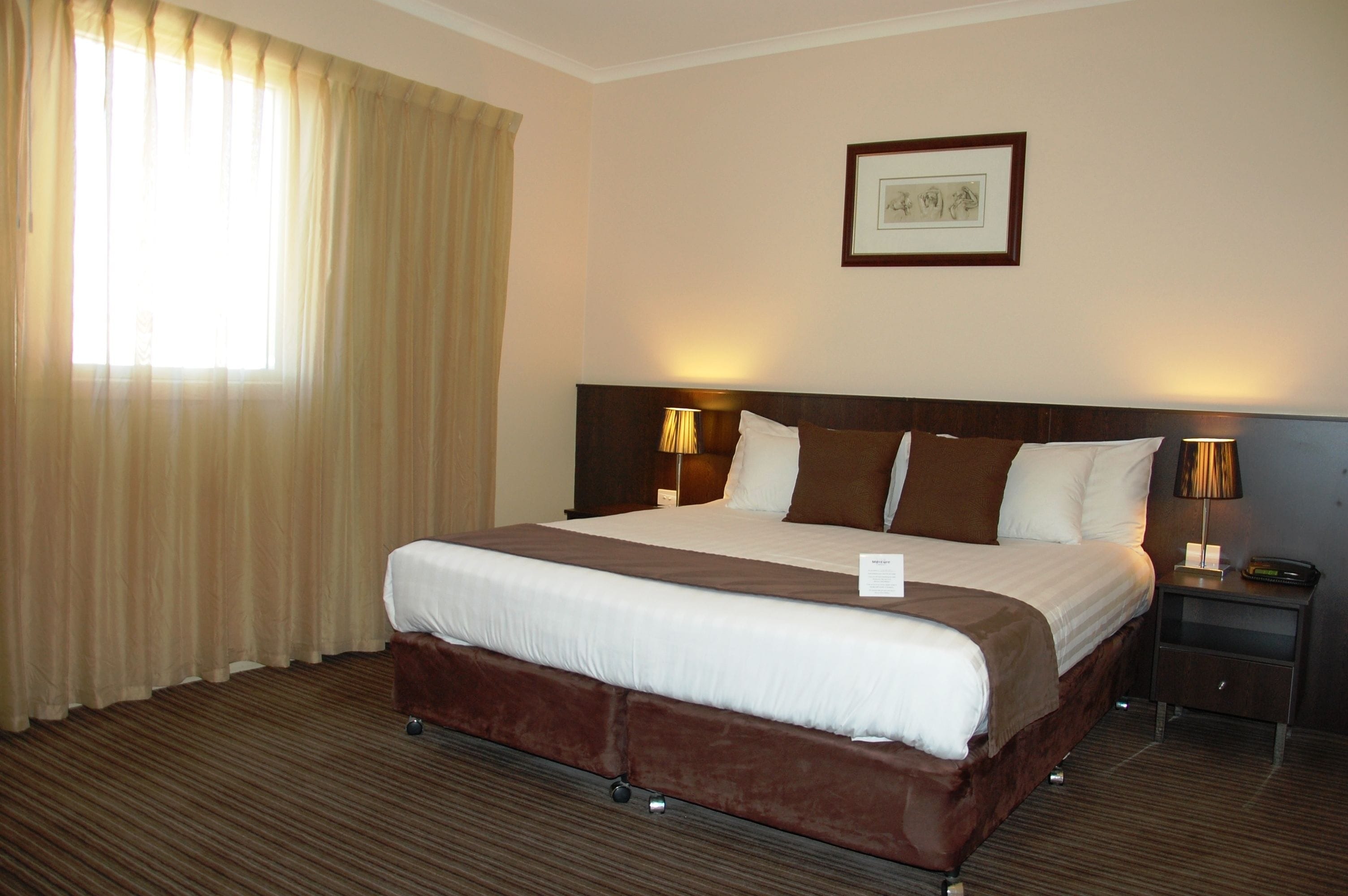 A hotel room with a large bed, a headboard, two bedside tables with lamps, a framed picture on the wall, and a window with curtains.
