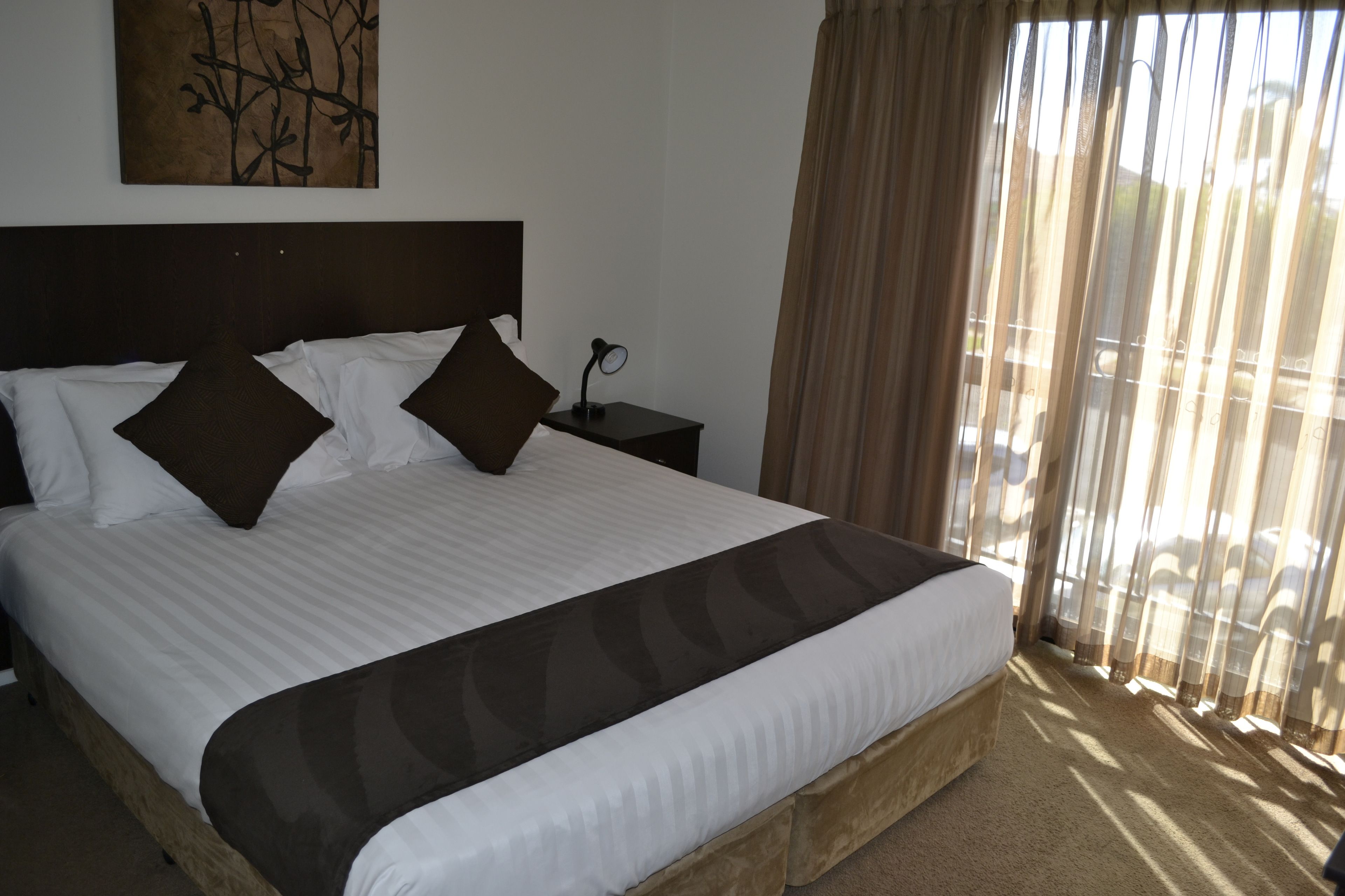 A neatly made bed with a dark headboard, white bedding, and two brown pillows. A small bedside table with a lamp is visible.