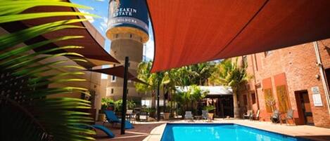 A swimming pool with a water tower in the background, surrounded by palm trees and a brick building.