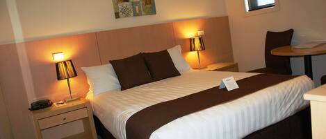 A neatly made bed with a brown and white striped bedspread, two bedside lamps, a small table with a chair, and a wall art piece above the bed.