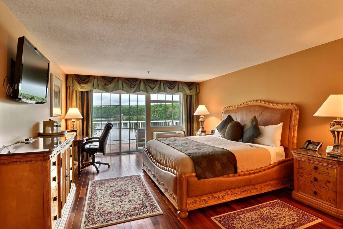 A bedroom with a large bed, a desk, a chair, a TV, and a balcony with a view of a lake.