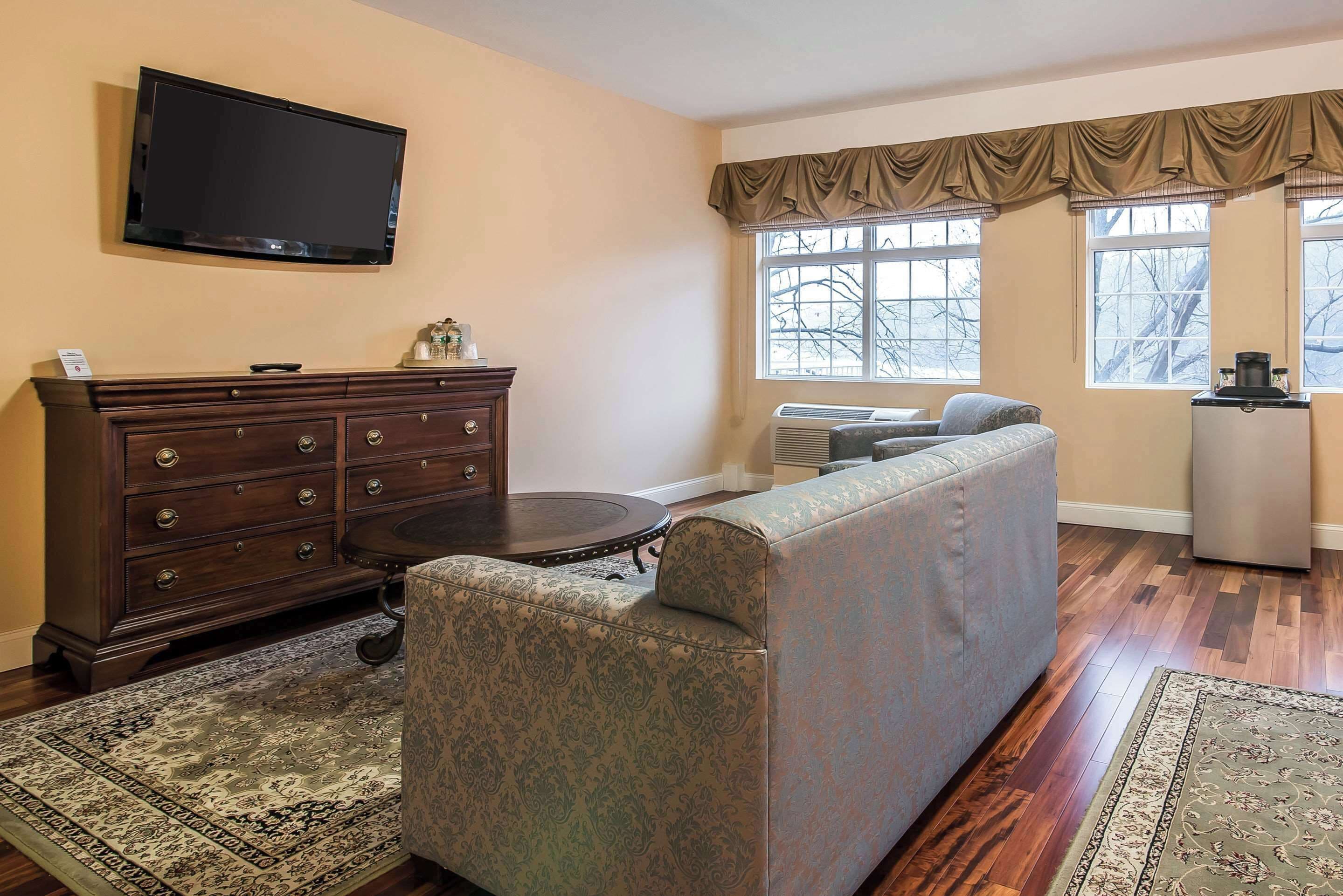 A living room with a dark wooden dresser, a flat-screen TV, a patterned armchair, a light-colored sofa, and a large window with curtains.