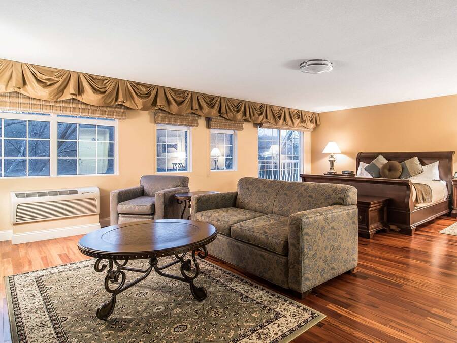 A living room with a sofa, armchair, and round table, a bedroom with a bed and nightstands, and large windows with curtains.