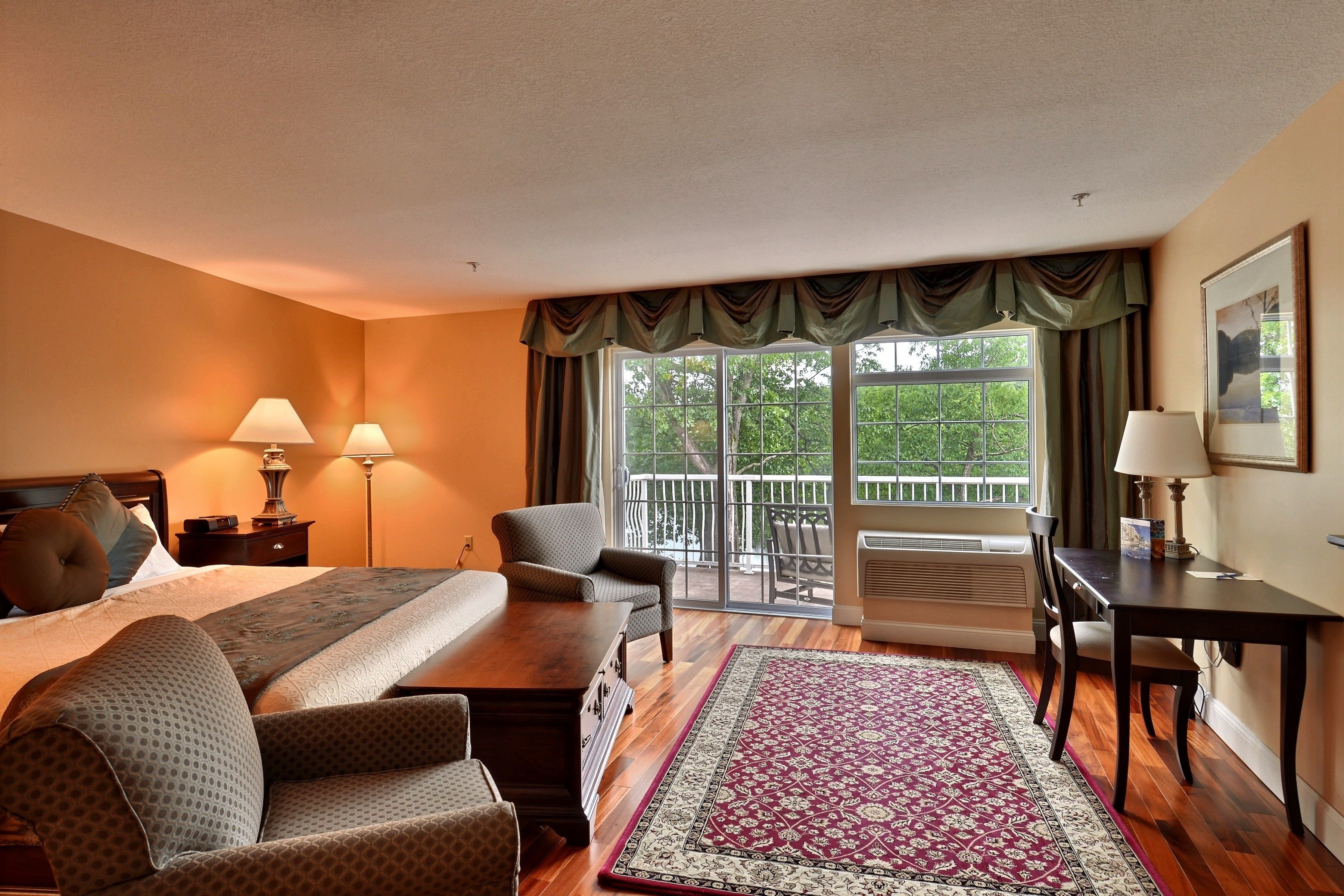 Habitación de hotel con cama, dos sillas, una mesa, escritorio, lámpara, ventana grande con cortinas y vistas a la vegetación.