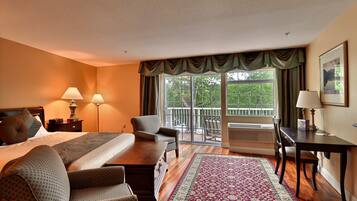 A hotel room with a bed, two chairs, a table, a desk, a lamp, a large window with curtains, and a view of greenery outside.