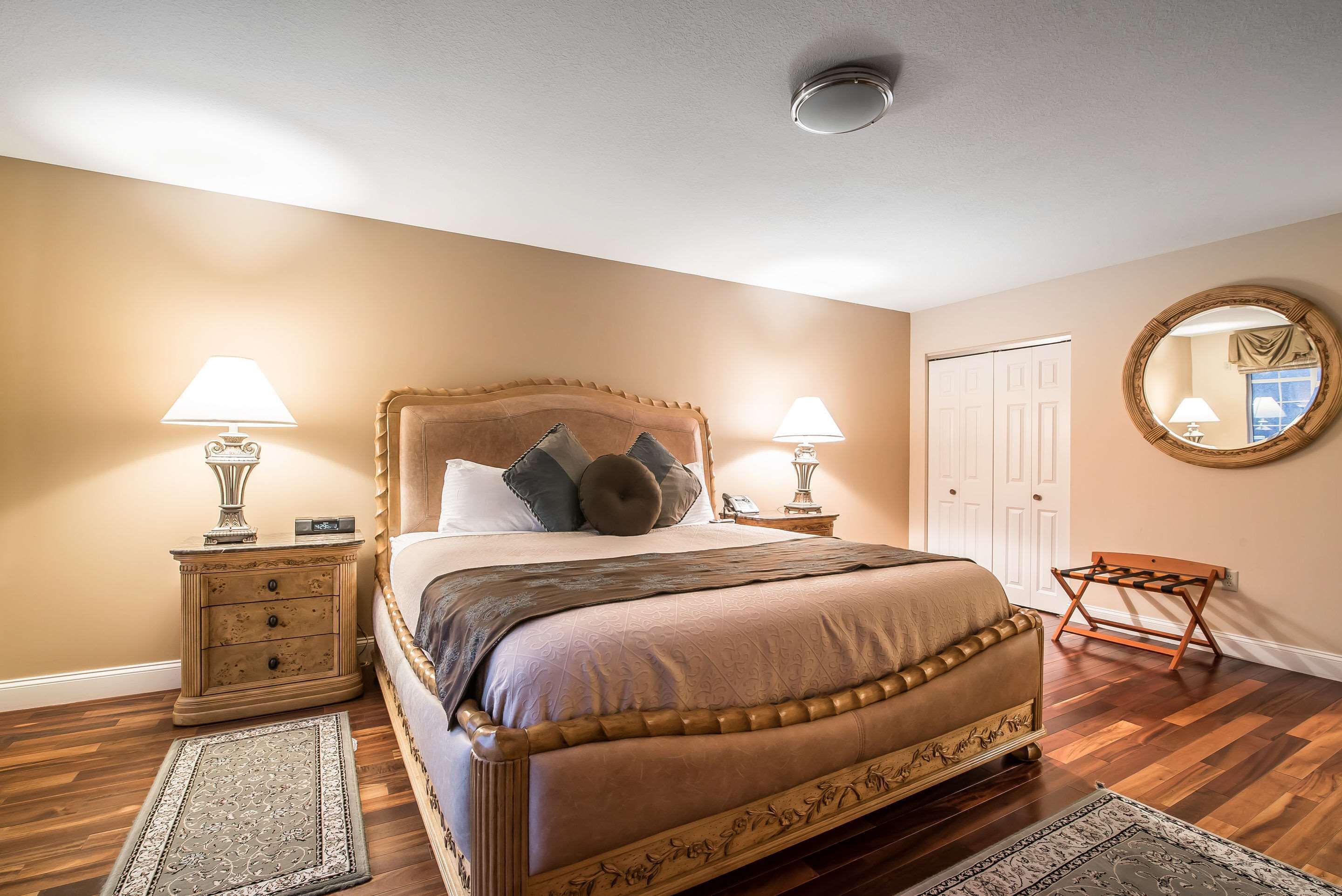 A bedroom with a large bed, two bedside tables with lamps, a wooden chair, a round mirror, and a door.