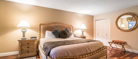 A bedroom with a large bed, two bedside tables with lamps, a wooden chair, a round mirror, and a door.