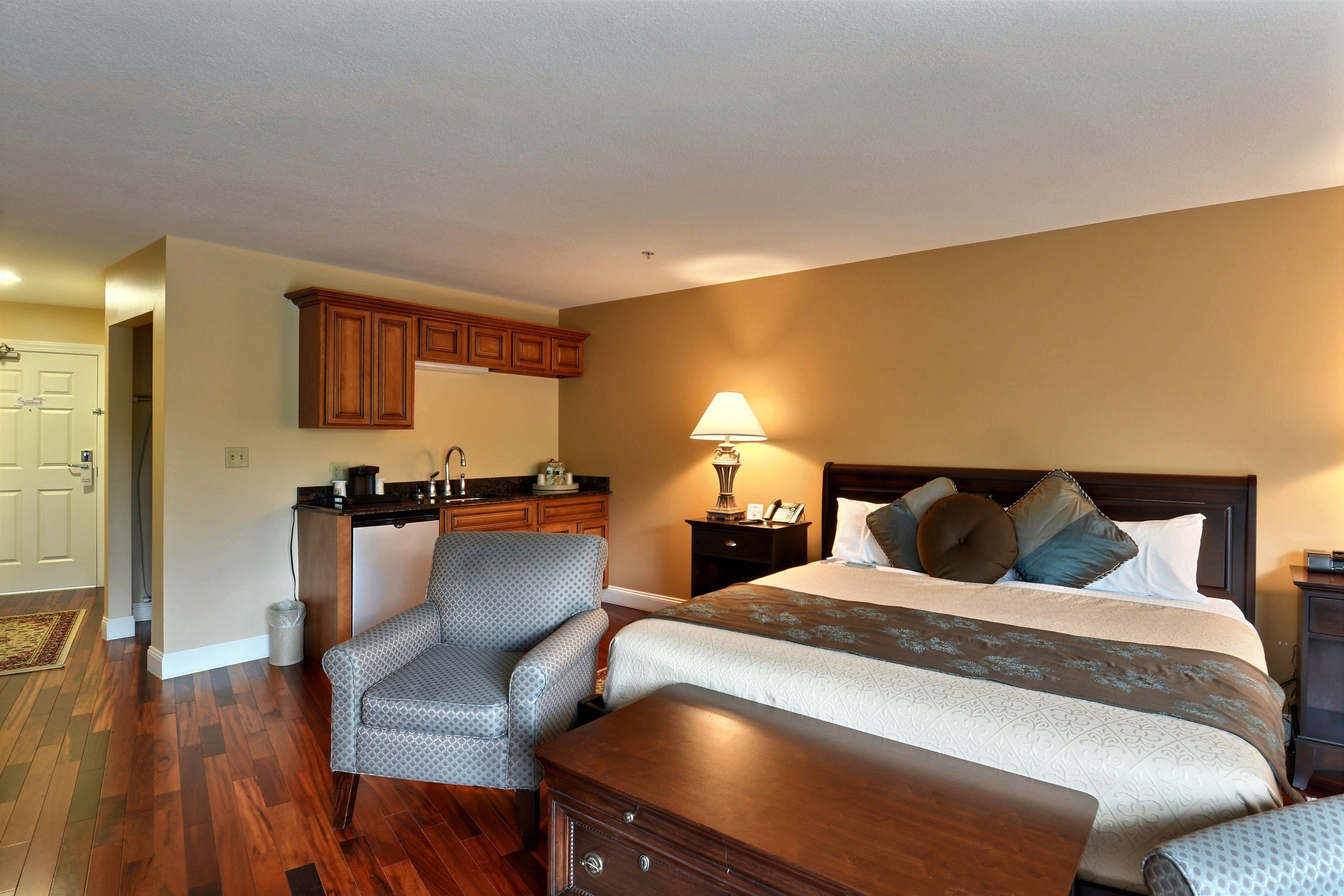 A hotel room with a bed, a chair, a desk, a lamp, and wooden cabinets.