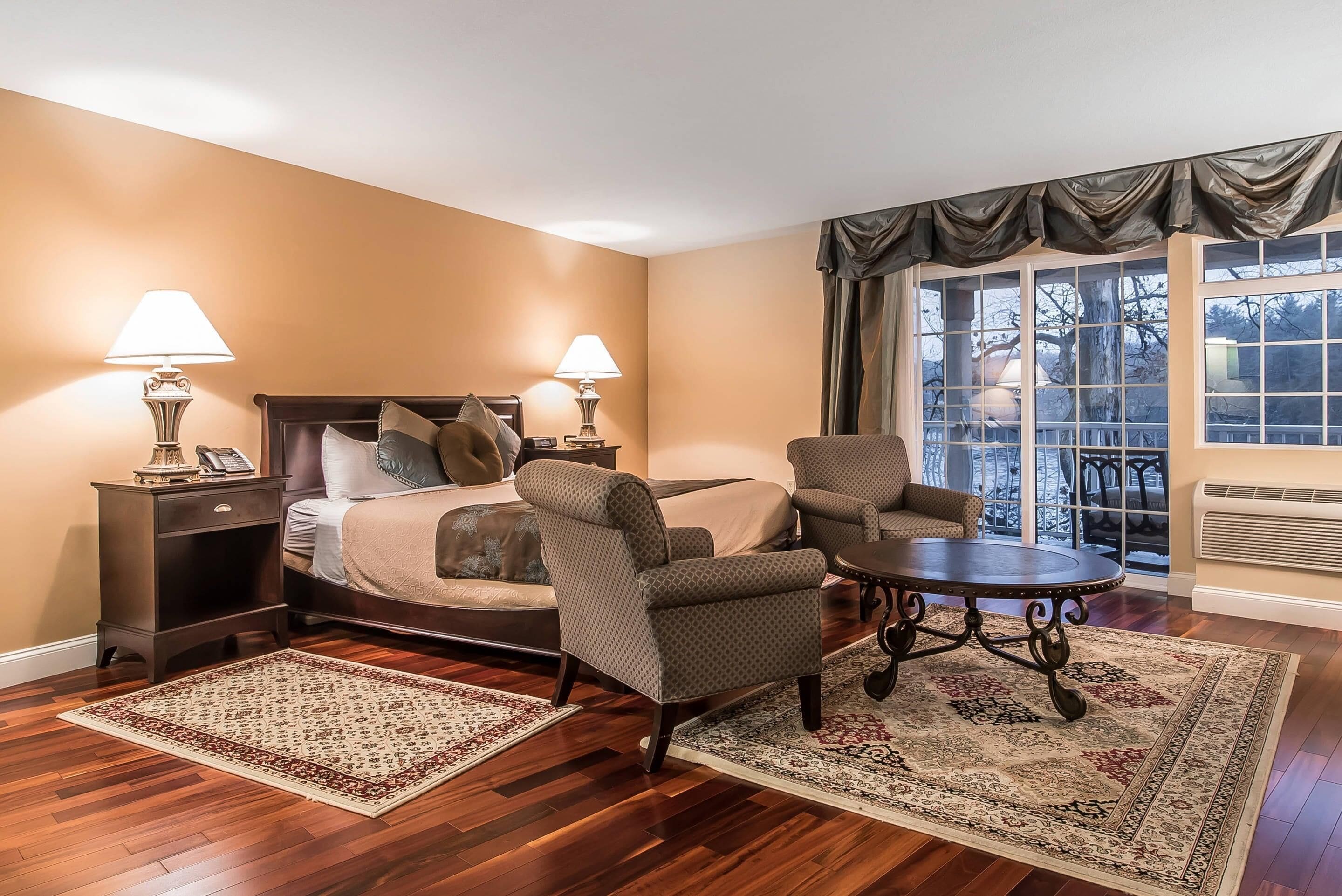 Quarto de hotel com uma cama, duas mesas de cabeceira com abajures, uma cadeira, uma mesa e uma janela grande com vista para árvores.