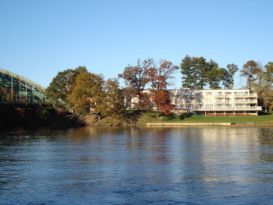 A building with a large balcony overlooking a body of water, a bridge in the foreground, and trees with autumn foliage.