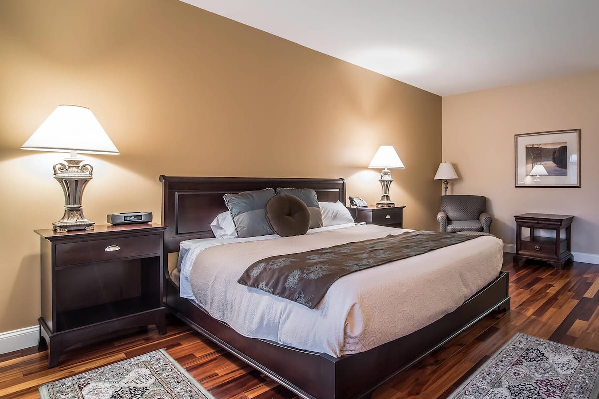A neatly arranged bedroom with a large bed, two bedside tables with lamps, a chair, and a framed picture on the wall.