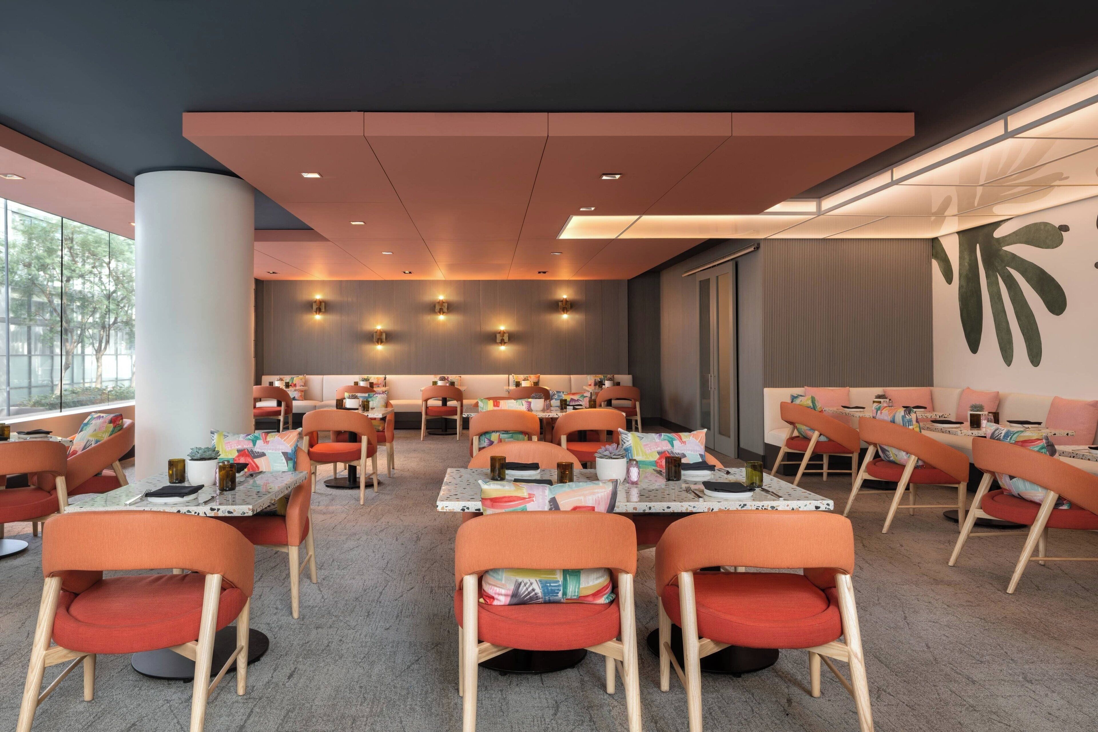 Un espace de restauration moderne doté de plusieurs tables et chaises, avec un éclairage combinant des sources naturelles et artificielles, et une grande fenêtre offrant une vue sur l’extérieur.