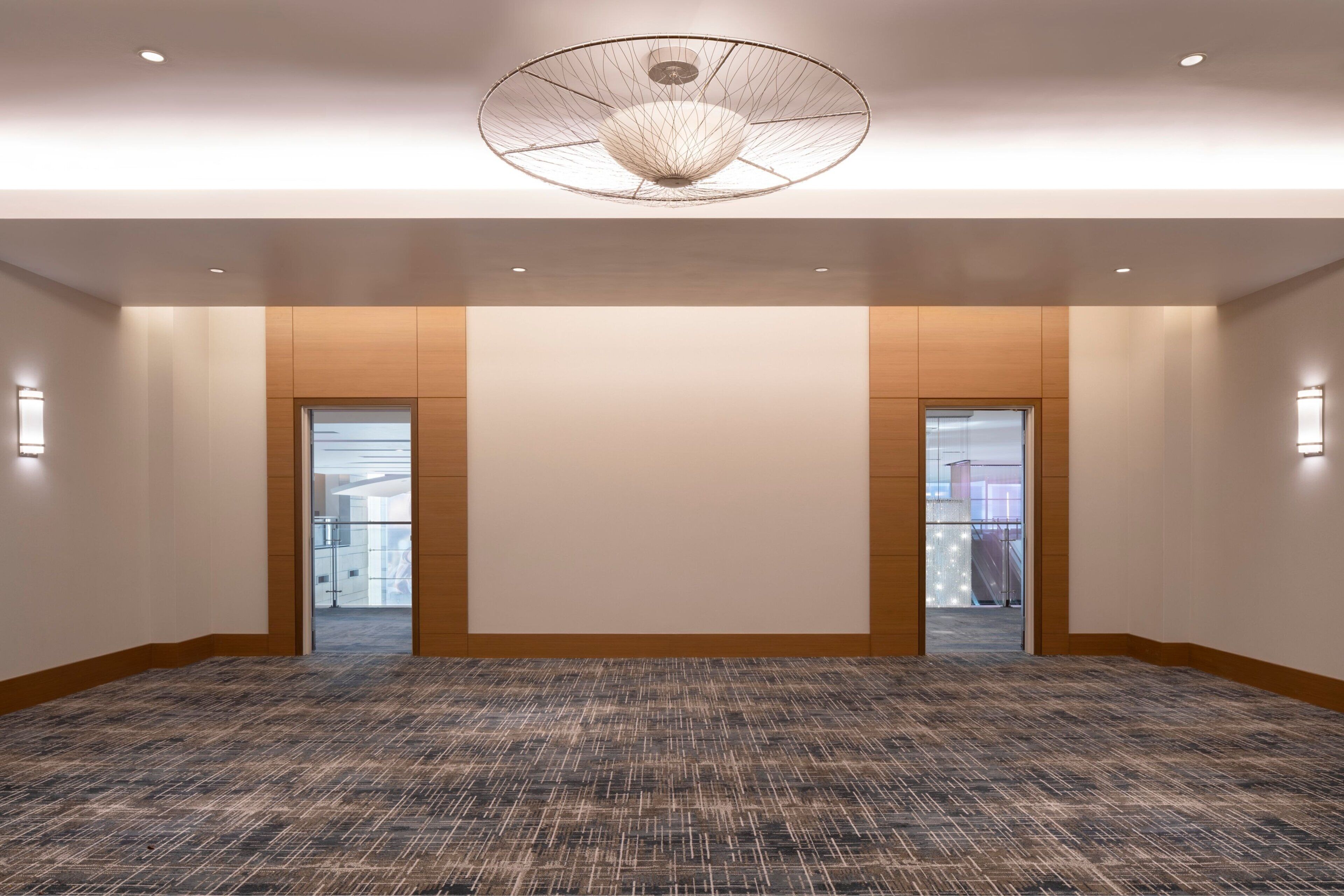 A spacious, modern hotel lobby with a patterned carpet, a large circular ceiling light, and wooden paneling on the walls.