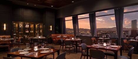 A restaurant with multiple tables and chairs, large windows with curtains, and a city view.