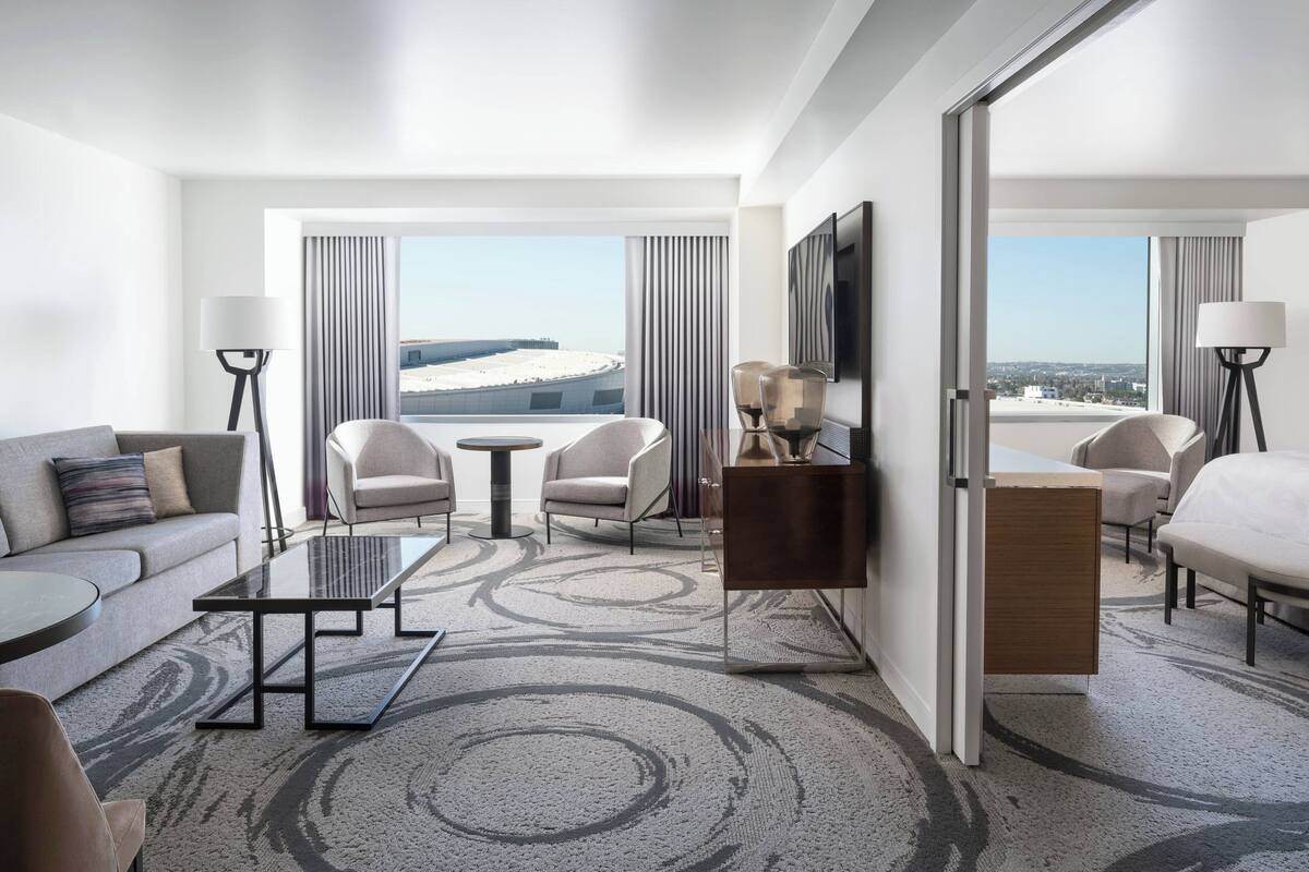 A modern hotel room with a sofa, two armchairs, a coffee table, a TV, and a view of a stadium.