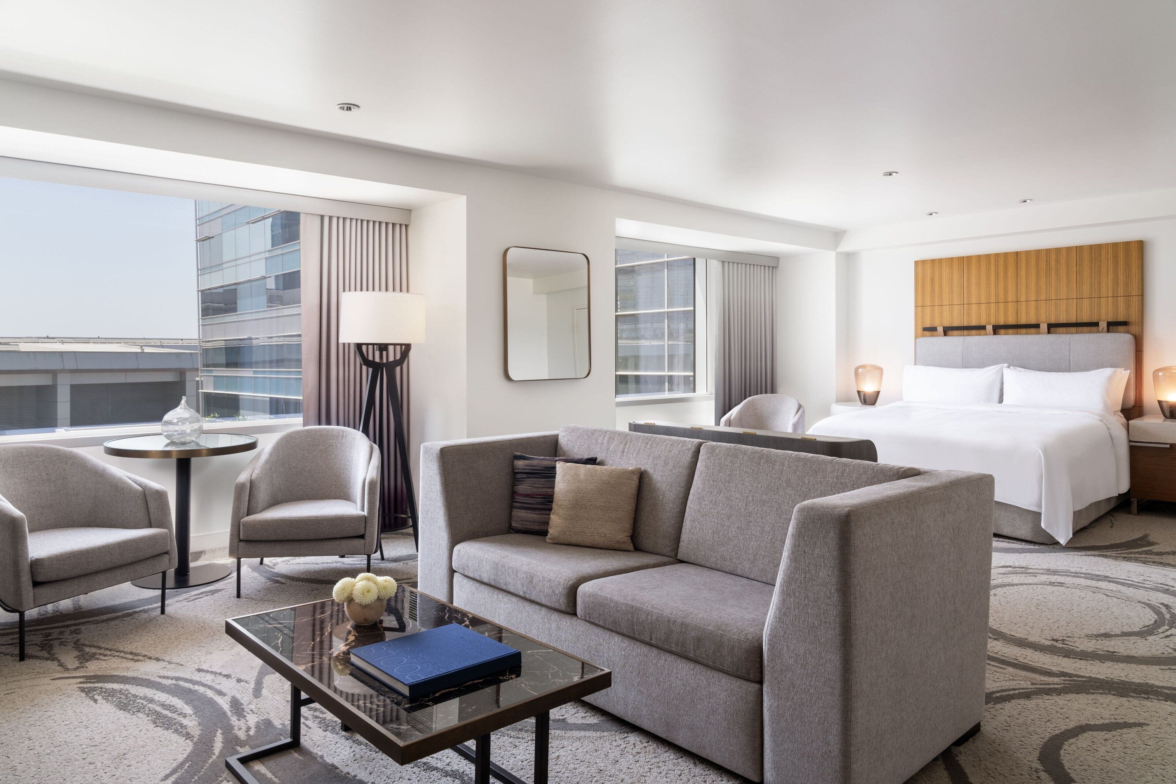 A modern hotel room with a sofa, a small table with a book, a bed, and a view of the city.