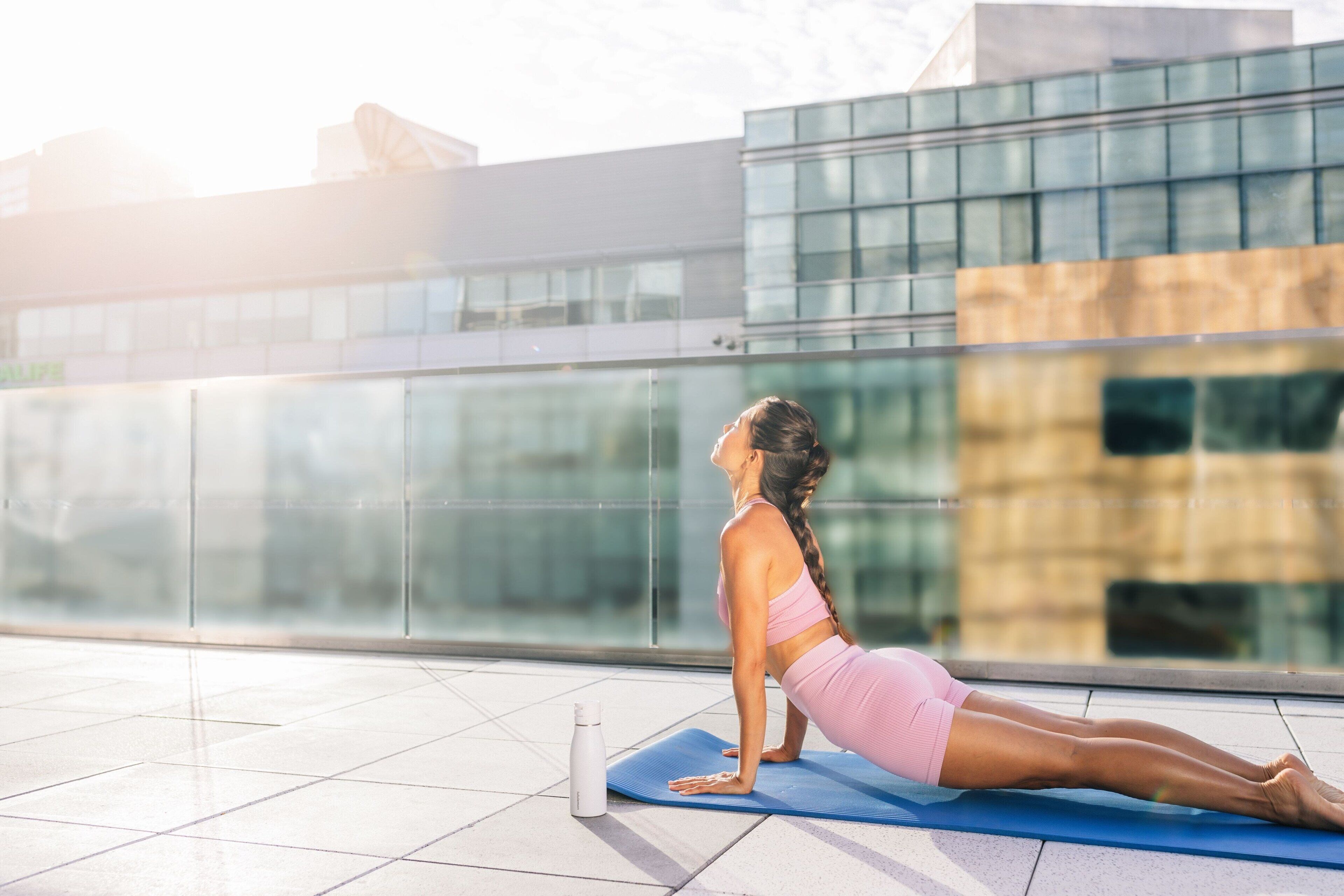 Una persona realizando una postura de yoga sobre una esterilla azul al aire libre, con un edificio moderno de fondo.