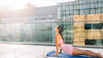 Una persona realizando una postura de yoga sobre una esterilla azul al aire libre, con un edificio moderno de fondo.