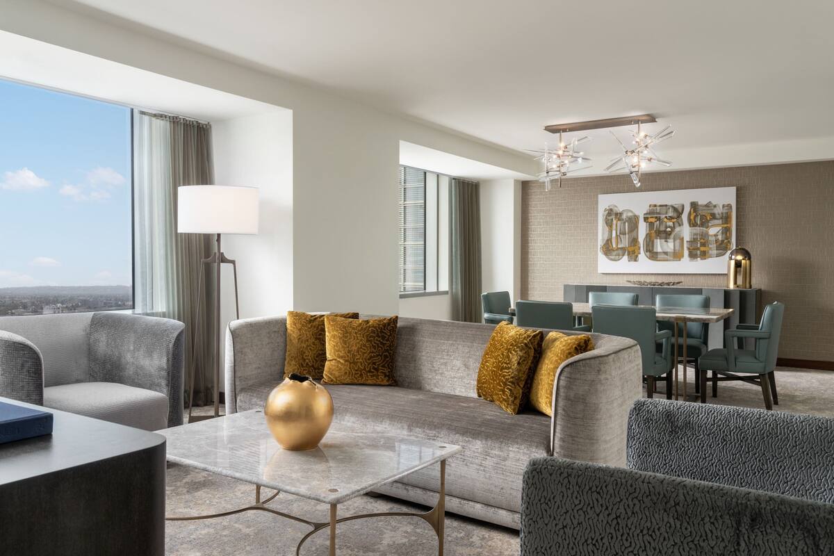 A modern hotel room with a sofa, a coffee table, a dining area, and a large window offering a city view.