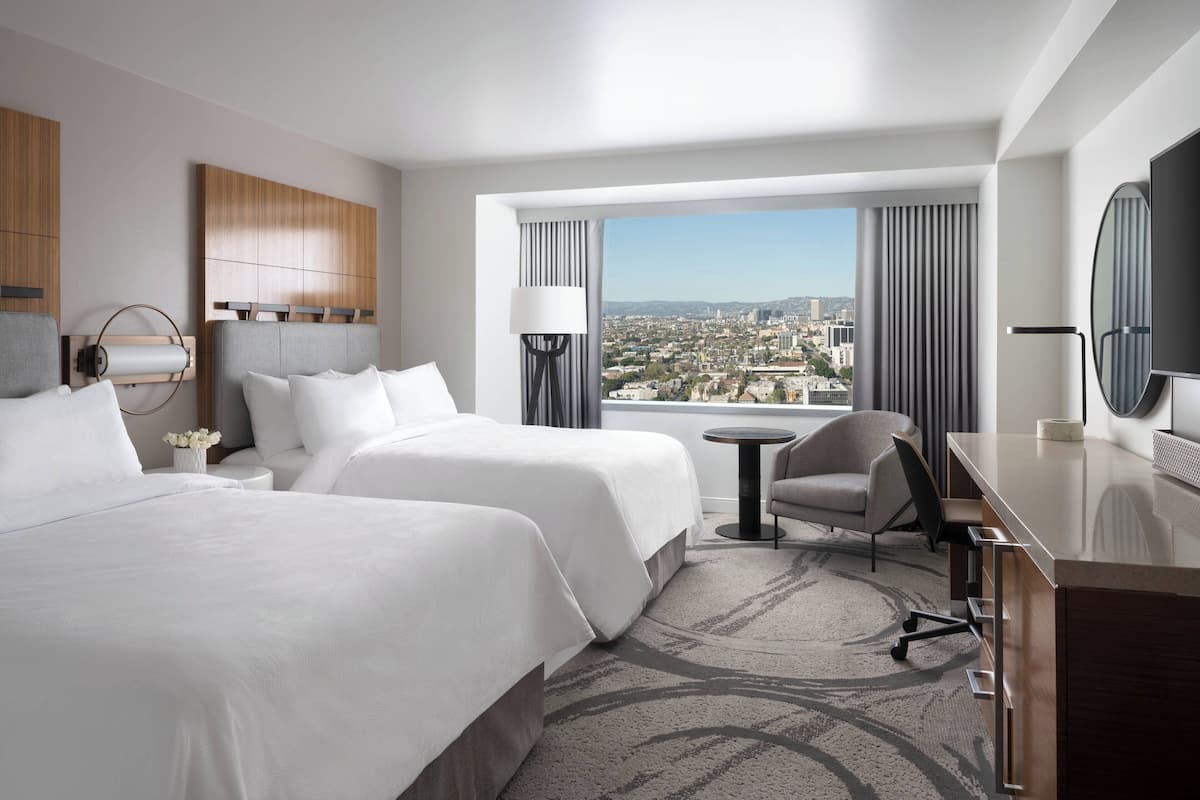 A hotel room with two beds, a desk, a chair, a TV, and a large window with a city view.