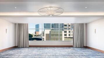 A large, empty room with a patterned carpet, a large window with curtains, and a modern ceiling light fixture.