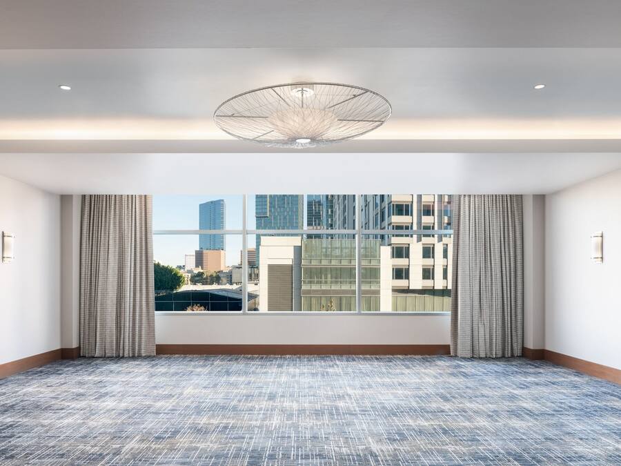 A large, empty room with a patterned carpet, a large window with curtains, and a modern ceiling light fixture.