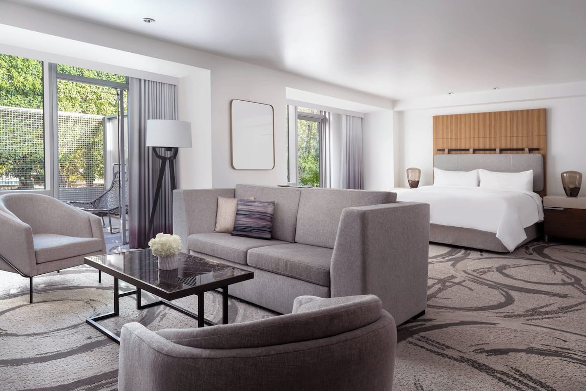 A modern hotel room with a large bed, a sofa, a coffee table, and a view of greenery outside.