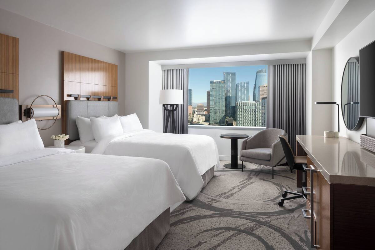 A hotel room with two beds, a desk, a chair, a TV, and a view of the city skyline.