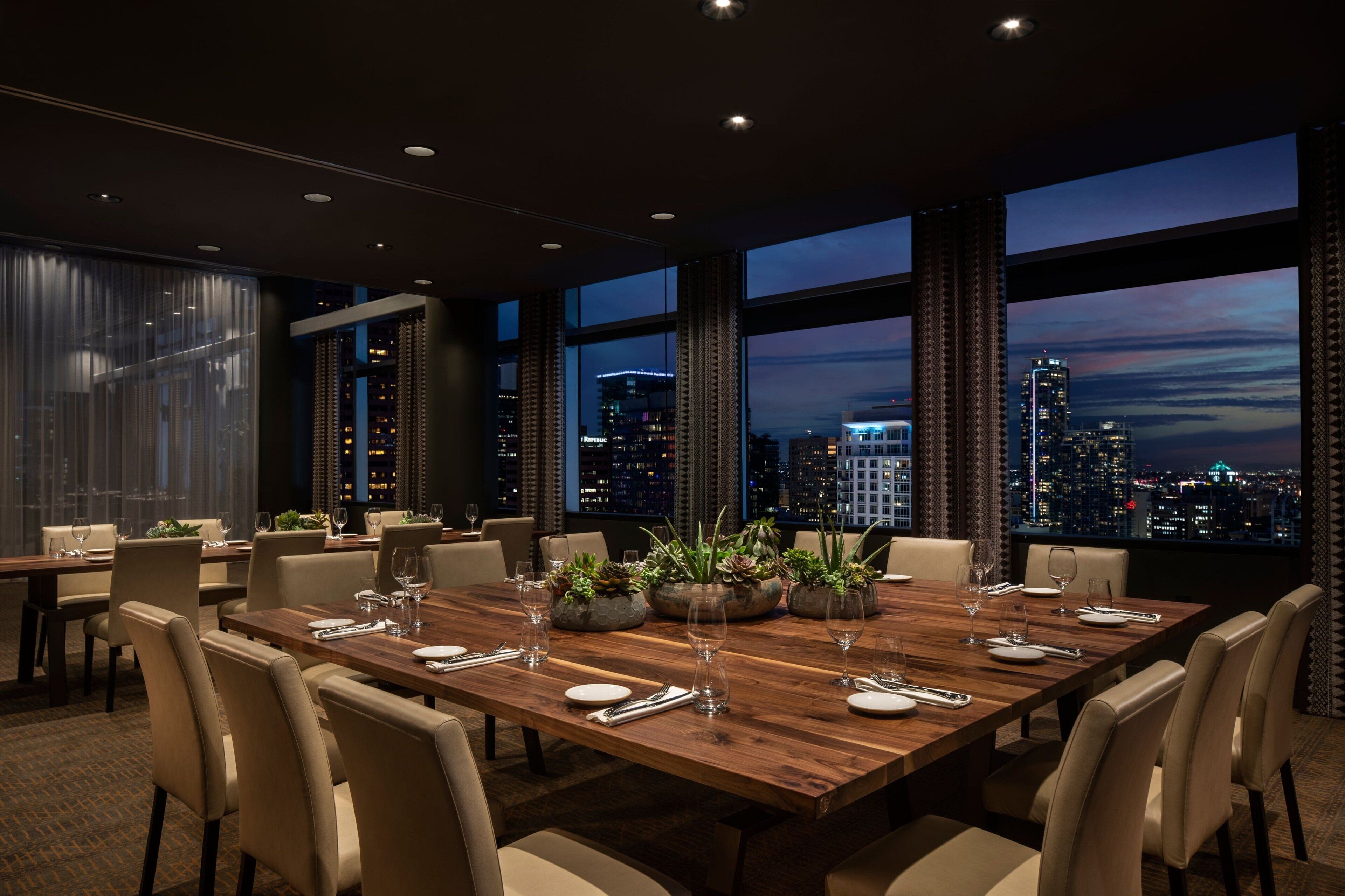Comedor con una gran mesa de madera dispuesta para seis personas, rodeada de sillas y vistas panorámicas de la ciudad a través de amplios ventanales.