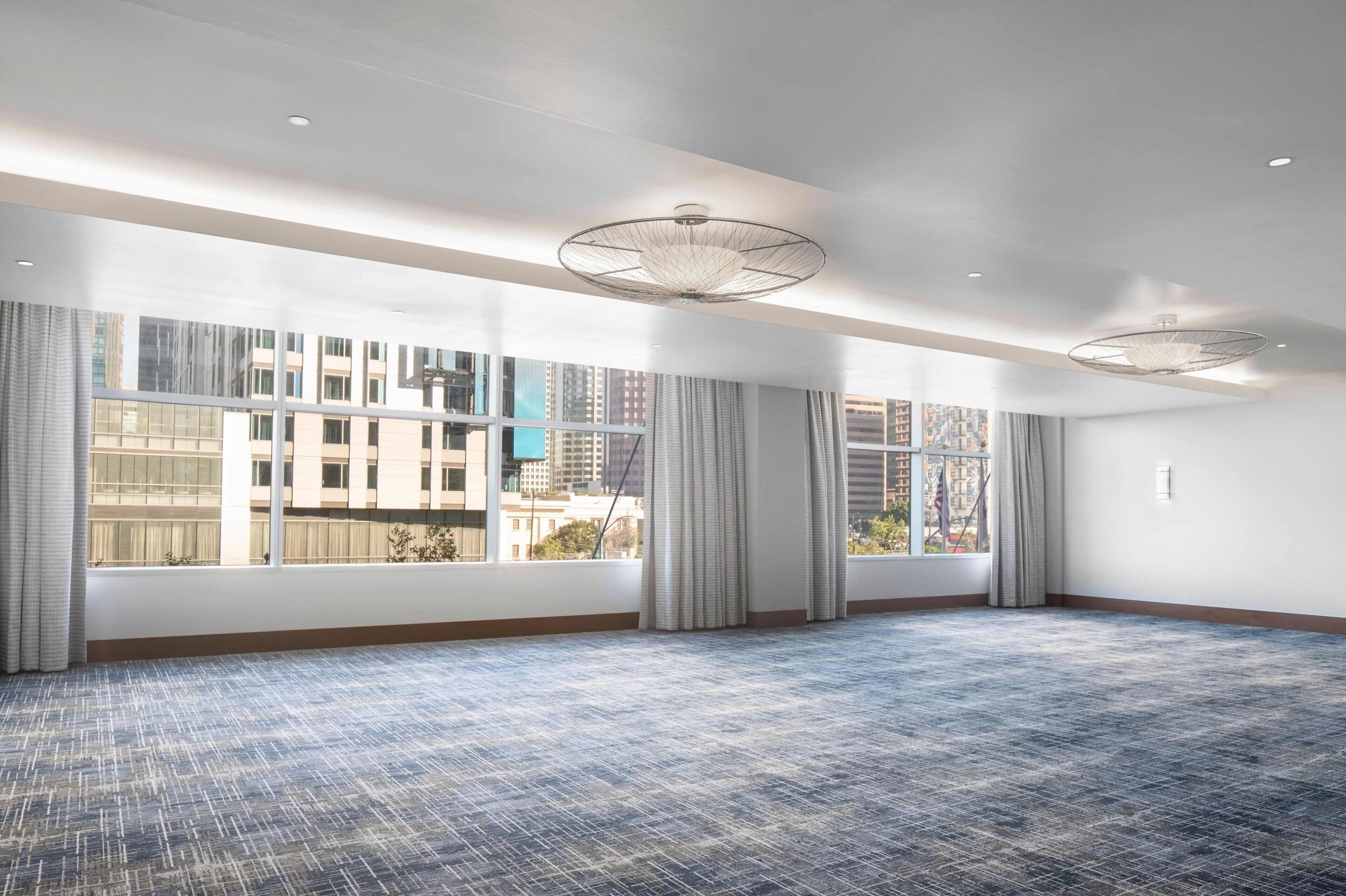 A large, empty room with a patterned carpet, large windows with curtains, and a modern ceiling light fixture.