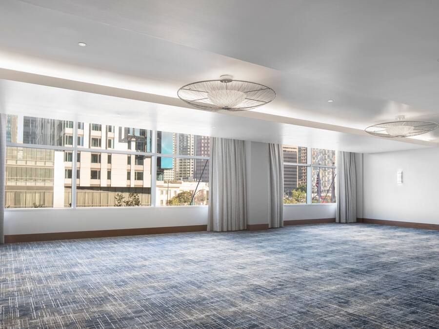 A large, empty room with a patterned carpet, large windows with curtains, and a modern ceiling light fixture.