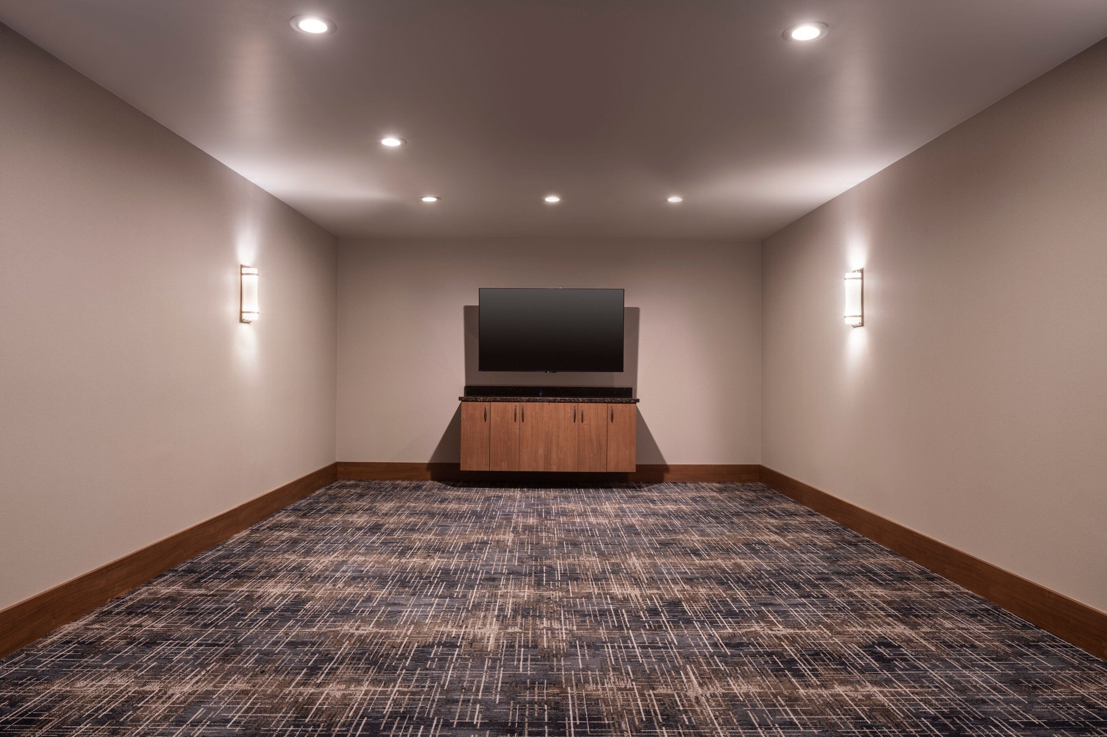 Una habitación con piso alfombrado, una televisión de pantalla plana sobre un atril de madera y luces empotradas en el techo.