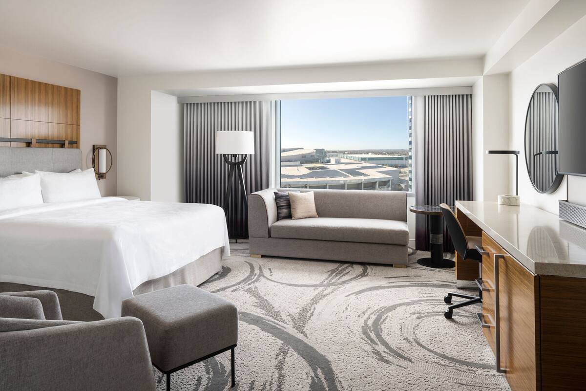 A hotel room with a large bed, a sofa, a desk, and a TV. There is a view of the city through the window.