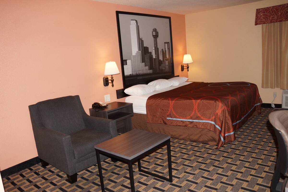 Habitación de hotel con cama, dos lámparas de noche, una silla, una mesita y una obra de arte con vista urbana en la pared.