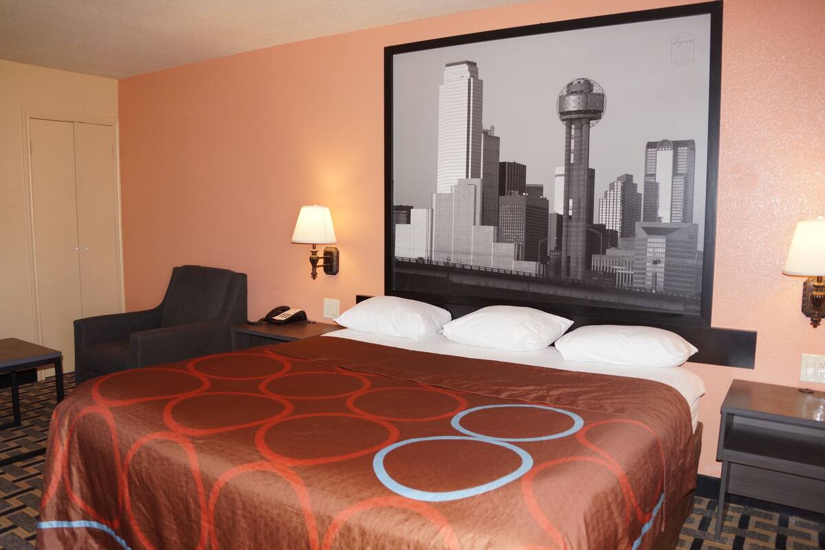 Habitación de hotel con una cama grande, dos mesitas de noche, una silla, un escritorio y un cuadro con una vista urbana en la pared.