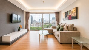 A modern living room with a sofa, a glass coffee table, a TV mounted on the wall, and a city view through large windows. - Olive Hotel & Residence Bangkok Sukhumvit 101 near True Digital Park (Bangkok)