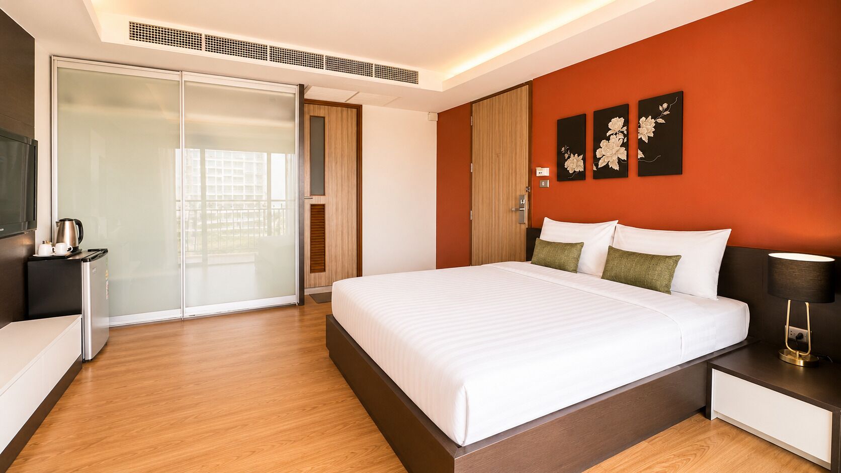 Une chambre d’hôtel moderne avec un grand lit, une table de chevet avec une lampe, une armoire et une porte-fenêtre coulissante donnant sur un balcon.