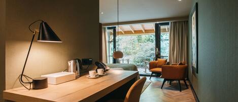 A modern room with a wooden desk, a lamp, a cup, a kettle, and a chair. There is a window with curtains and a view of greenery outside.