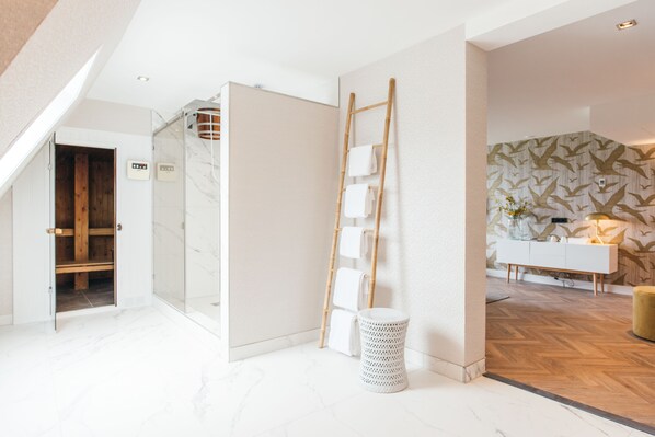 A modern hotel room with a glass shower enclosure, a wooden ladder, a white trash bin, and a patterned wallpaper wall.